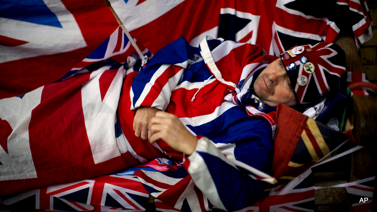 Royal fan John Loughrey, aged 60, with his Union flag designed outfit and flags demonstrates how he has laid on a bench to sleep for the last four nights across the street from the Lindo Wing of St Mary's Hospital in London, Thursday, April 23, 2015. Britain's Kate the Duchess of Cambridge is expected to give birth to her second child with her husband Prince William at the hospital in the coming days or weeks.  A small number of dedicated royal fans are waiting or camping outside the hospital awaiting the imminent birth. (AP Photo/Matt Dunham)
