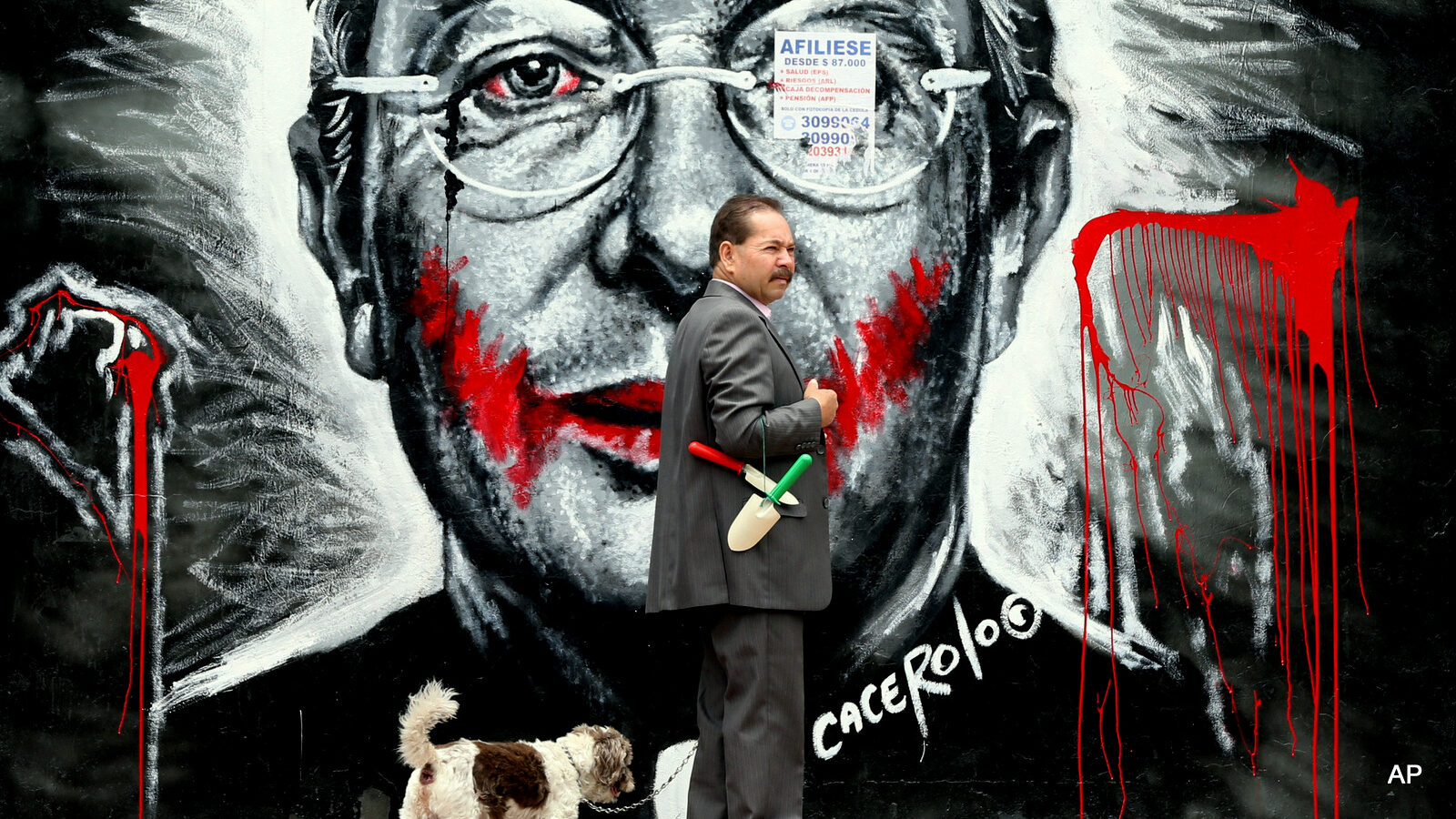 In this March 18, 2015 photo, a man walks his dog next to a mural depicting former President Alvaro Uribe, in Bogota, Colombia. Bogota’s laissez-faire attitude toward graffiti contrasts with that in many other Latin American cities. Buenos Aires, Argentina, last year raised penalties for street art, which it considers to be vandalism. In the Peruvian capital of Lima, the mayor this month had city workers cover up several down murals by graffiti artists.