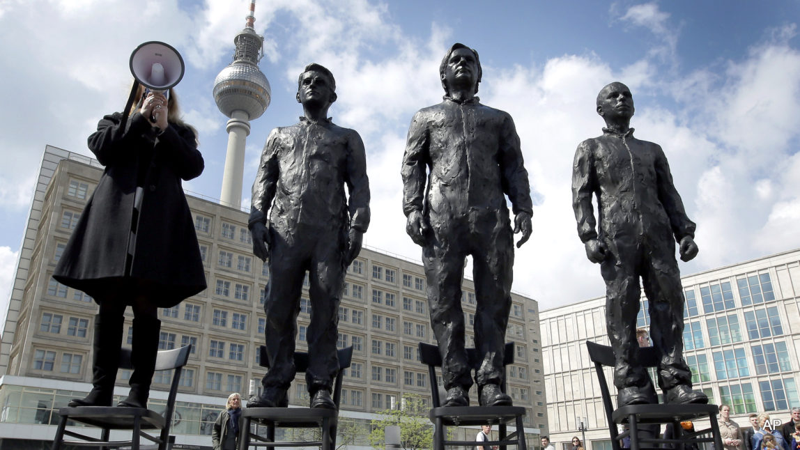 A women delivers a speech as she stands on a chair of the public art project "Anything to Say?" at the Alexander Square in Berlin, Germany, Friday, May 1, 2015. The sculpture of the Italian artist Davide Dormino shows the whistleblowers Chelsea Manning, Julian Assange and Edward Snowden, from right, to honour their courage.