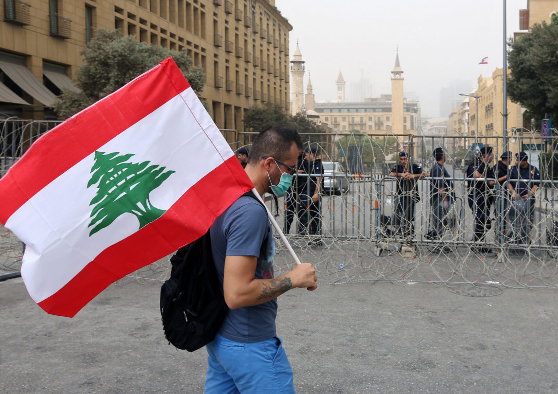 A Lebanese anti-government protester holds his national flag as Lebanese riot policemen stand guard behind barbed wire that blocks a road leading to the parliament building, during a protest against the on-going trash crisis and government corruption, in downtown Beirut, Lebanon, Wednesday, Sept. 9, 2015. Lebanon's prime minister says he hopes that political talks between senior politicians will help end government paralysis that has sparked angry street protests. (AP Photo/Bilal Hussein)