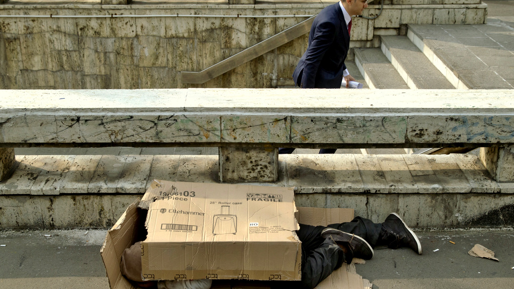 A pedestrian exits a subway station as a homeless man sleeps covered in cardboard packaging, outside the main railway station in Bucharest, Romania, Wednesday, Oct. 7, 2015. The sewers around the railway station are a popular shelter for the town's many homeless, especially in the bitter Balkan winter weather. (AP Photo/Vadim Ghirda)