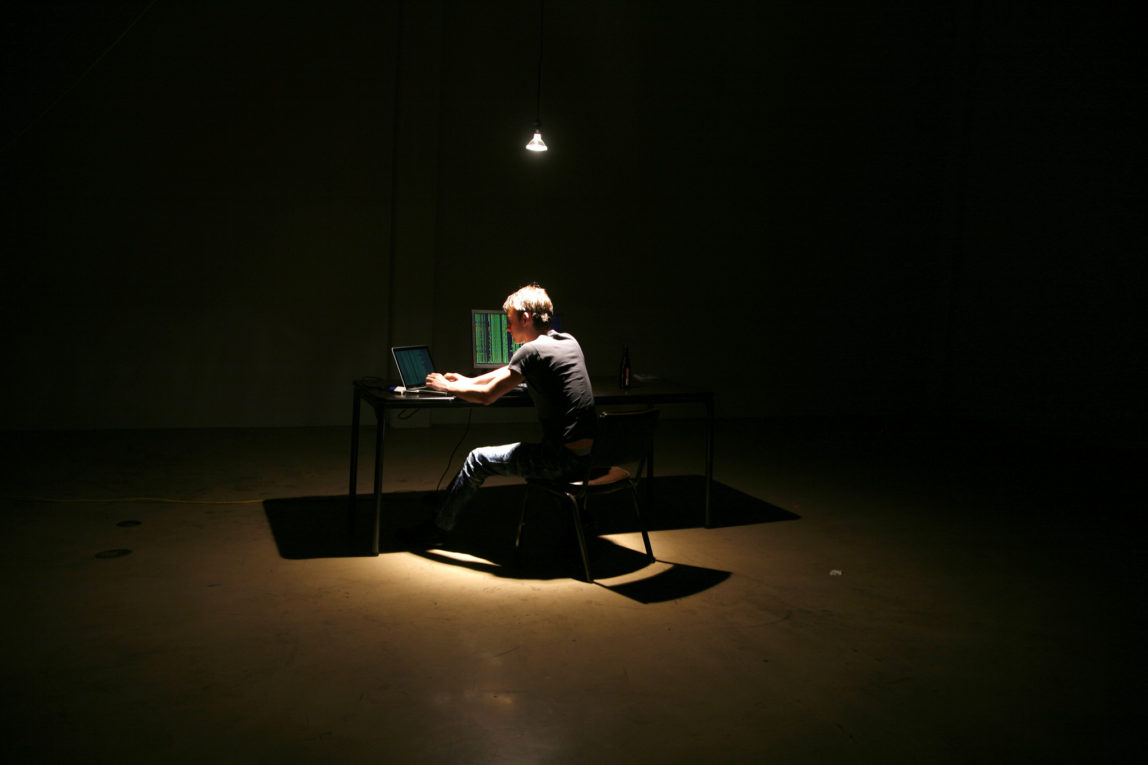 Alone in a warehouse, a hacker sits at a laptop while another screen displays data in green text. (Flickr / Brian Klug)