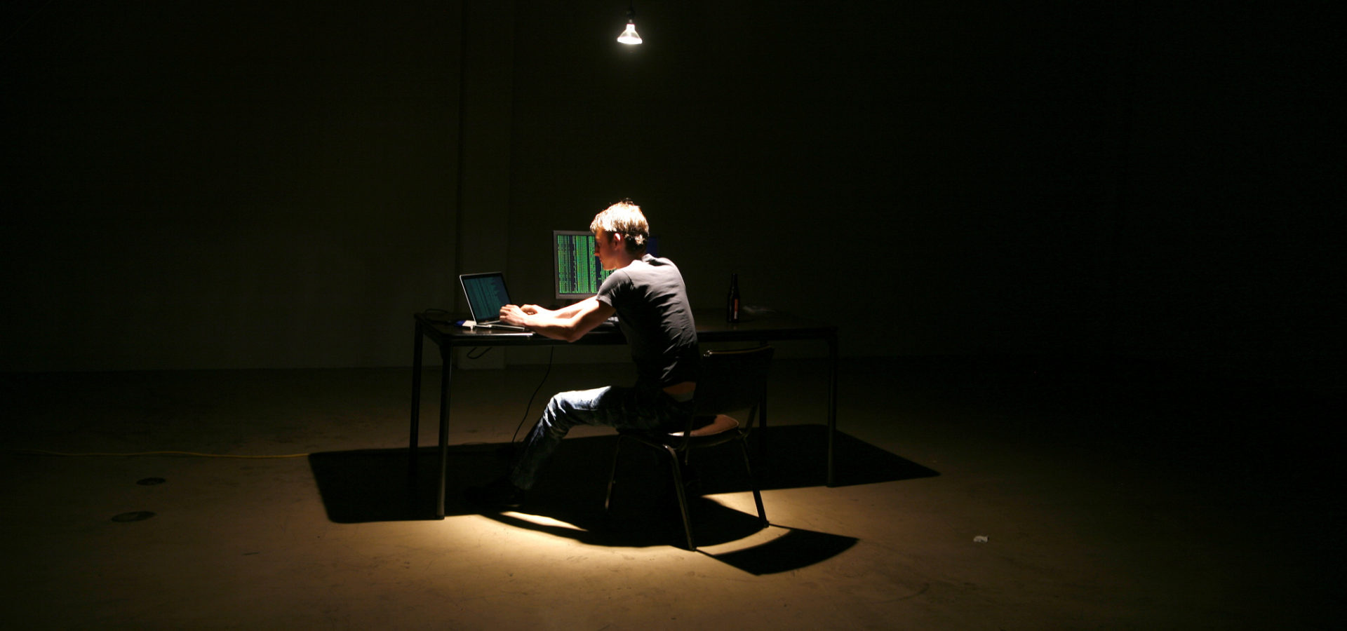 Alone in a warehouse, a hacker sits at a laptop while another screen displays data in green text. (Flickr / Brian Klug)