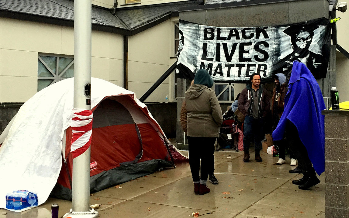 A group of protesters gather at the front entrance to the 4th Police Precinct station, Monday, Nov. 16, 2015, in Minneapolis, after a man was shot by Minneapolis police early Sunday morning. Community members and activists on Monday at a news conference demanded that police release the name of the officer involved as well as any video of the incident. (AP Photo/Kyle Potter)