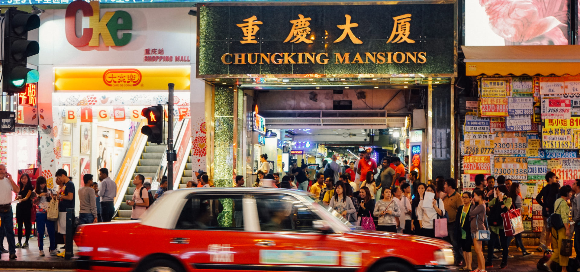 A red taxi drives past tthe busy exterior of the Chungking Mansions in Hong Kong. These squalid, overcrowded towers house many of the territory's 10,000 asylum seekers. (Shadowproof / Zachary Senn)