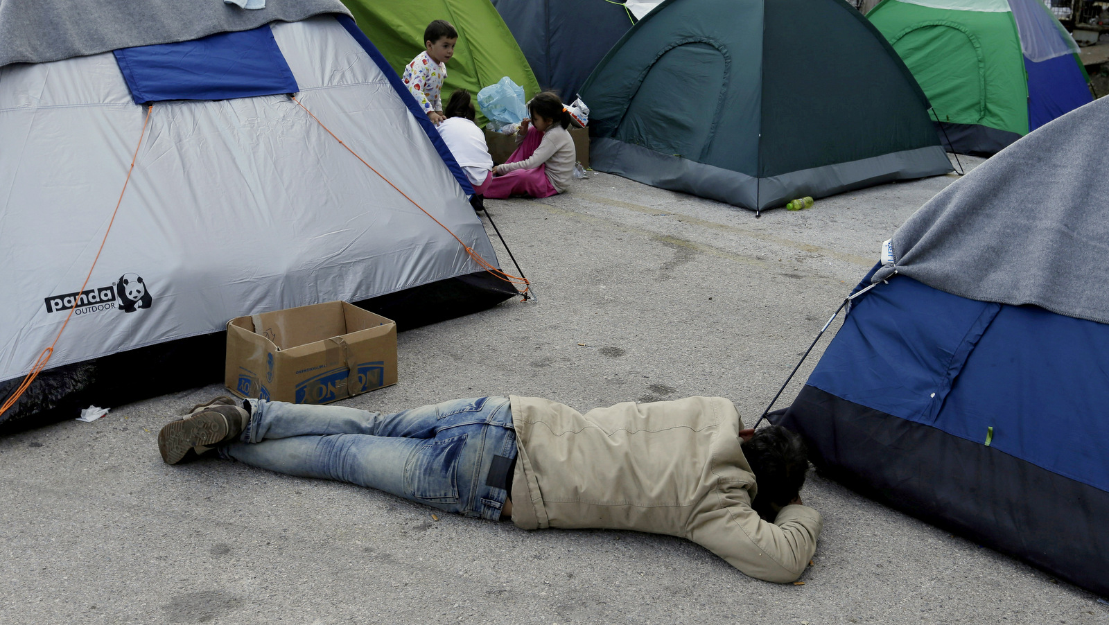 A man sleeps on the ground as children play outside their tents at the Athens' port of Piraeus, where over 5,500 refugees and migrants stay, on Monday, March 21, 2016. Greek Prime Minister Alexis Tsipras says an international agreement to limit the number of refugees and migrants traveling to Europe cannot be properly implemented unless smugglers on the Turkish coast are stopped. (AP Photo/Thanassis Stavrakis)