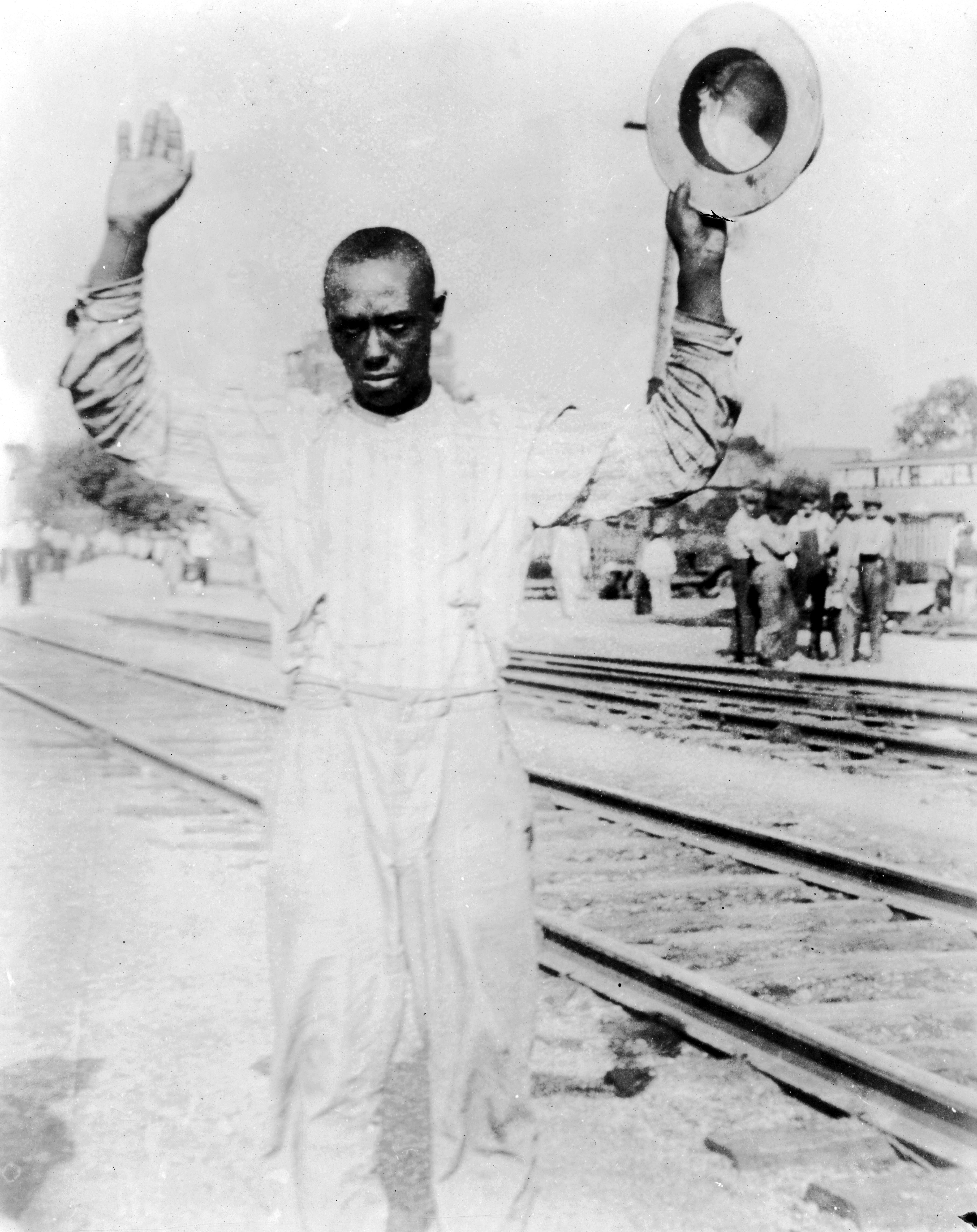 Photo of an African-American man being detained during the Tulsa Race Riot. The man is standing next to railroad tracks and is holding his hands in the air as if being arrested. Several white men watch from the other side of the tracks.