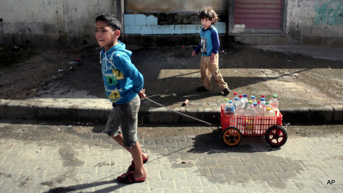 In this Saturday, April 16, 2016 photo, A Palestinian boy uses a homemade wagon made from a plastic crate to wheel bottles full of drinking water in front of a water supply station in Khan Younis refugee camp, southern Gaza Strip. Poor sewage treatment is a feature of life in Gaza, a result of infrastructure damaged during Israel's bombing campaigns and ongoing blockade.