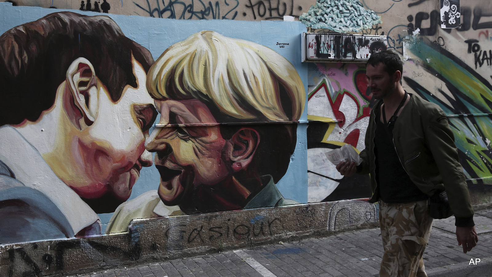 FILE - In this Sunday, Oct. 18, 2015 file photo, a man walks past street art depicting Greek Prime Minister Alexis Tsipras and German Chancellor Angela Merkel in Athens, Greece. Tsipras' decision to sign off on a bailout led to many in his left-wing Syriza party to quit in protest. 