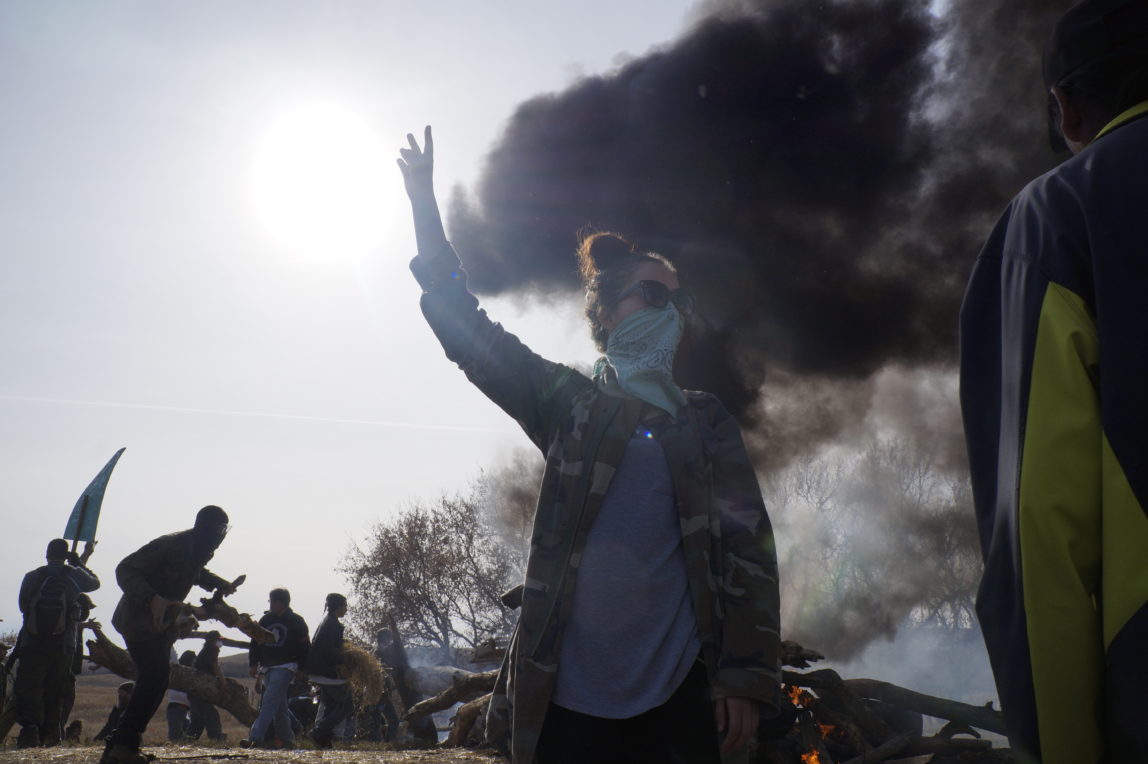 Gabriella Scarlett, a water protector from Canada, signals for peace as a fire barricade burns off County Road 134. Behind her, water protectors establish a fire barricade to hold police back from the site of construction of the Dakota Access pipeline. Oct. 27, 2016  (Derrick Broze for MintPress)