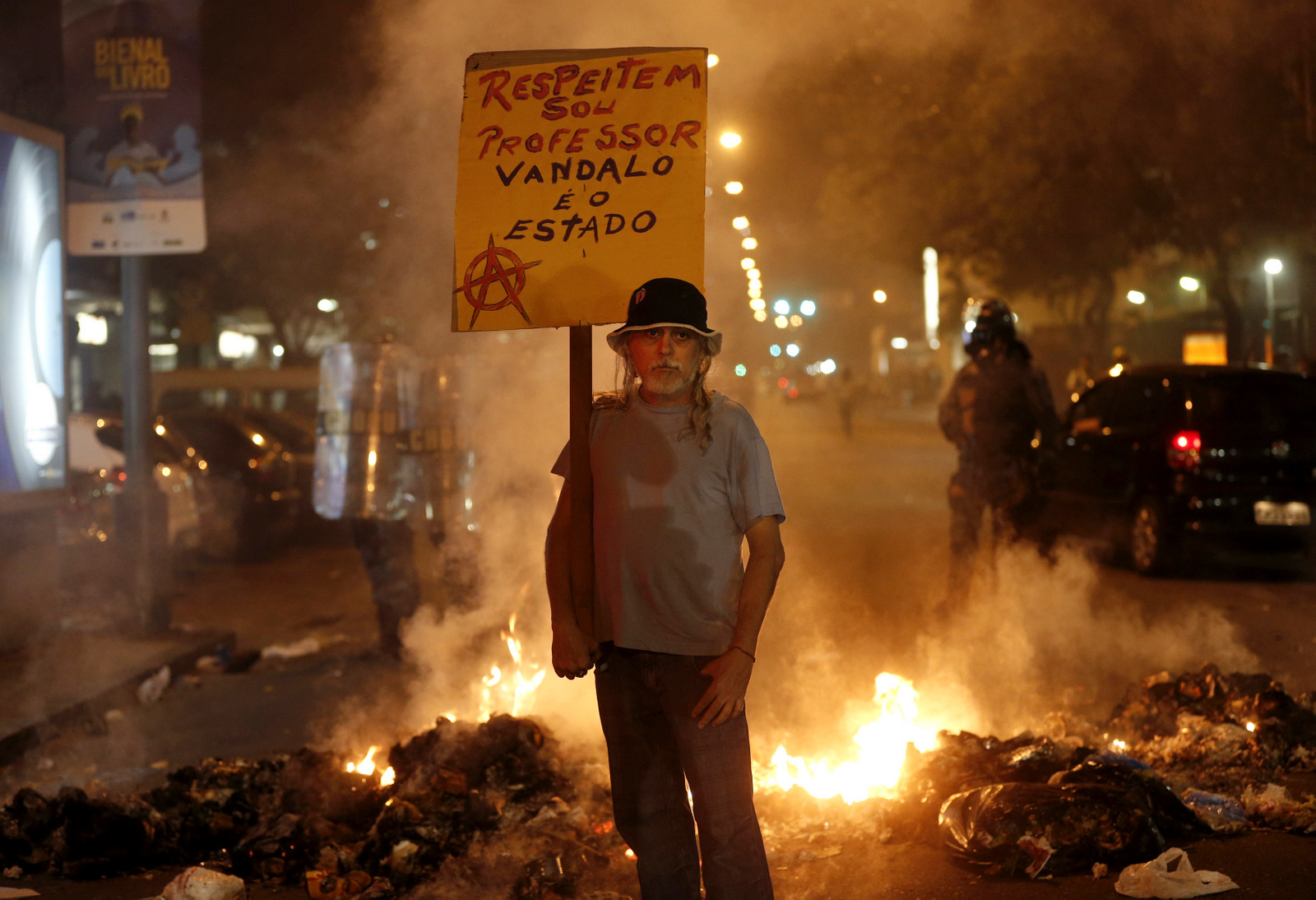 A man holds a sign that reads in Portuguese "Respect, I'm a teacher, the vandal is the state" at a burning barricade set up by protesters in Rio de Janeiro, Brazil. (AP/Silvia Izquierdo)