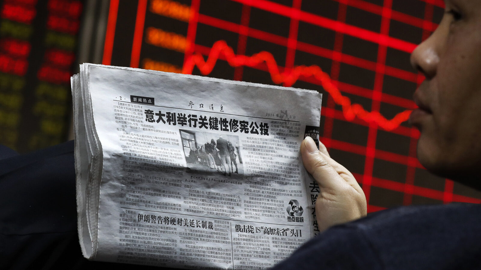 A man holds a newspaper as he looks at an electronic board displaying stock prices at a brokerage house in Beijing, Monday, Dec. 5, 2016. (AP/Andy Wong)