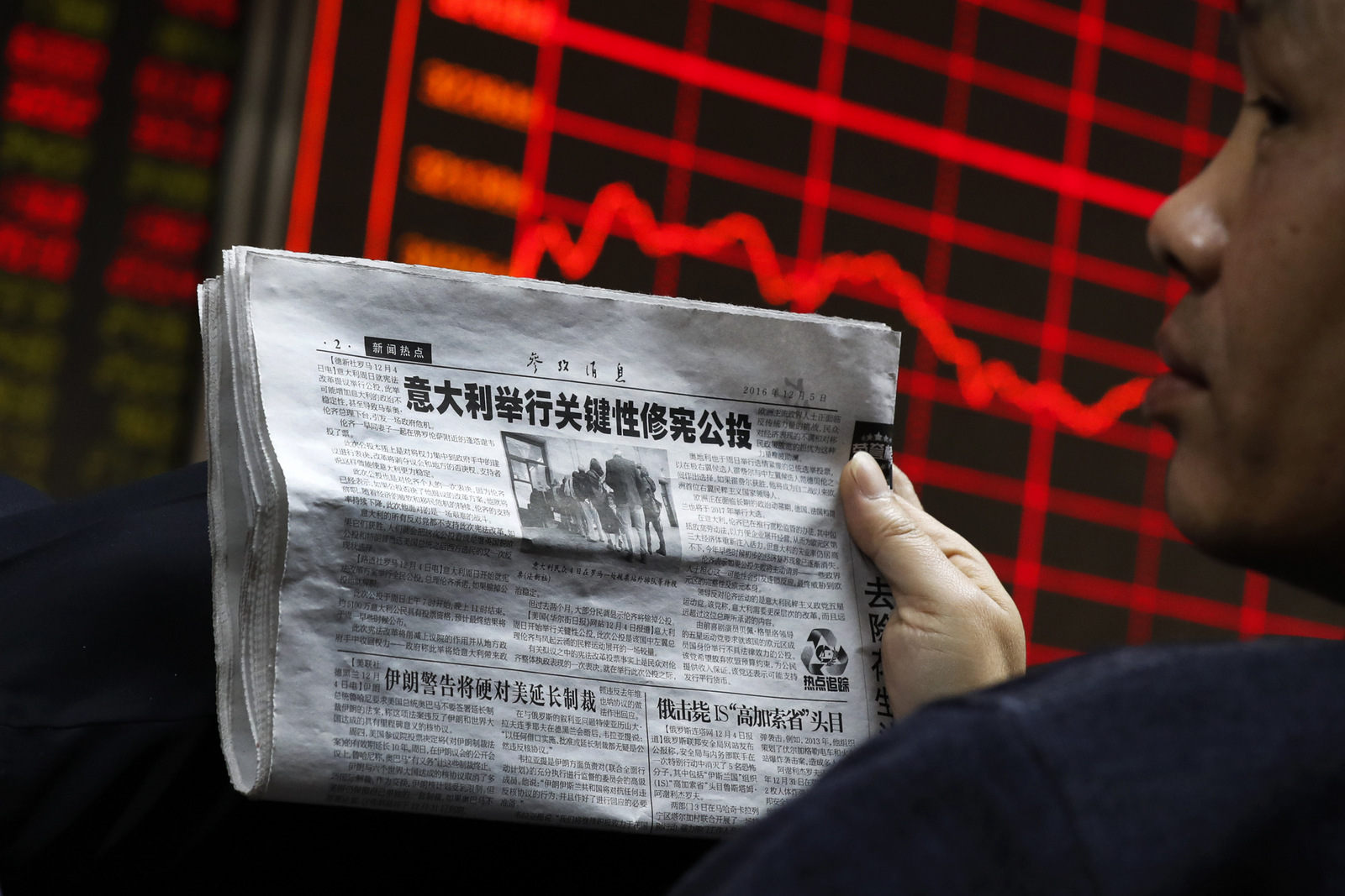 A man holds a newspaper  as he looks at an electronic board displaying stock prices at a brokerage house in Beijing, Monday, Dec. 5, 2016.  (AP/Andy Wong)