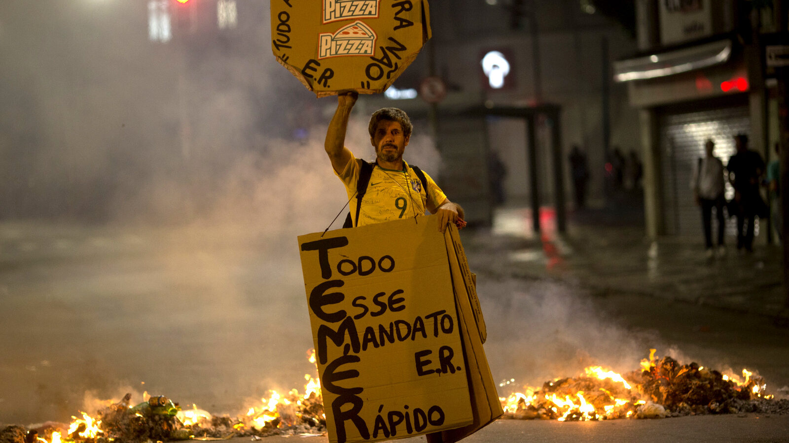 A demonstrator holds a sign against Brazil's President Michel Temer at a burning road block set up by protesters in Rio de Janeiro, Brazil, Thursday, May 18, 2017. Temer rejected calls for his resignation, saying he will fight allegations that he endorsed the paying of hush money to a former lawmaker jailed for corruption. The signs read in Portuguese "His entire mandate will be quick," and "Not this time. It all ends in pizza," meaning there will be consequences for Temer. (AP/Silvia Izquierdo)