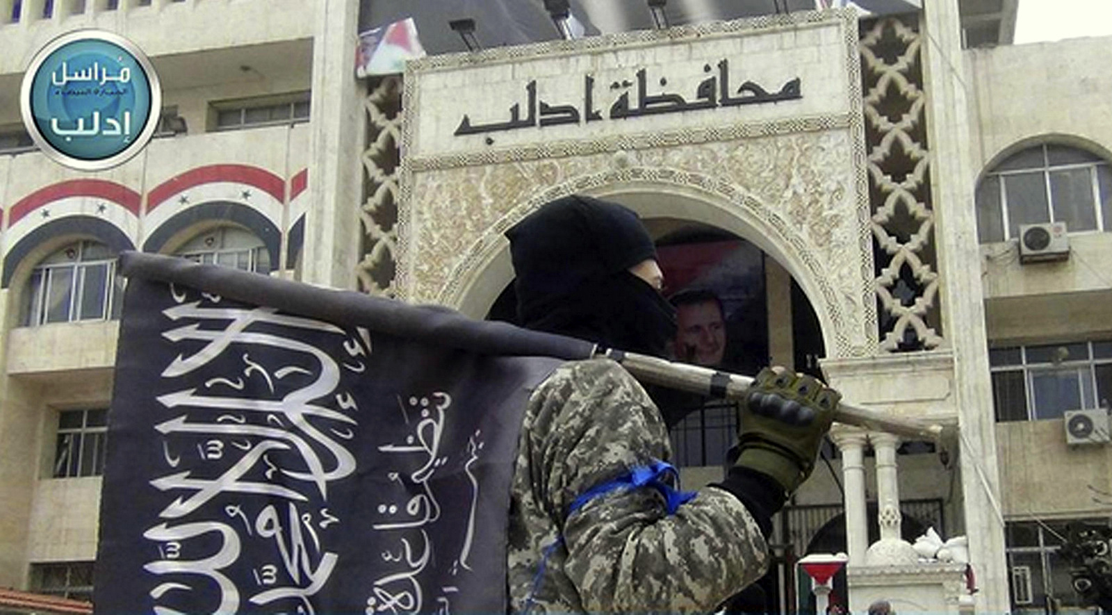 当他站在叙利亚北部伊德利布省的州长大楼前时，一名努斯拉战斗机举着他的团队旗帜。 （图片：Twitter通过AP）