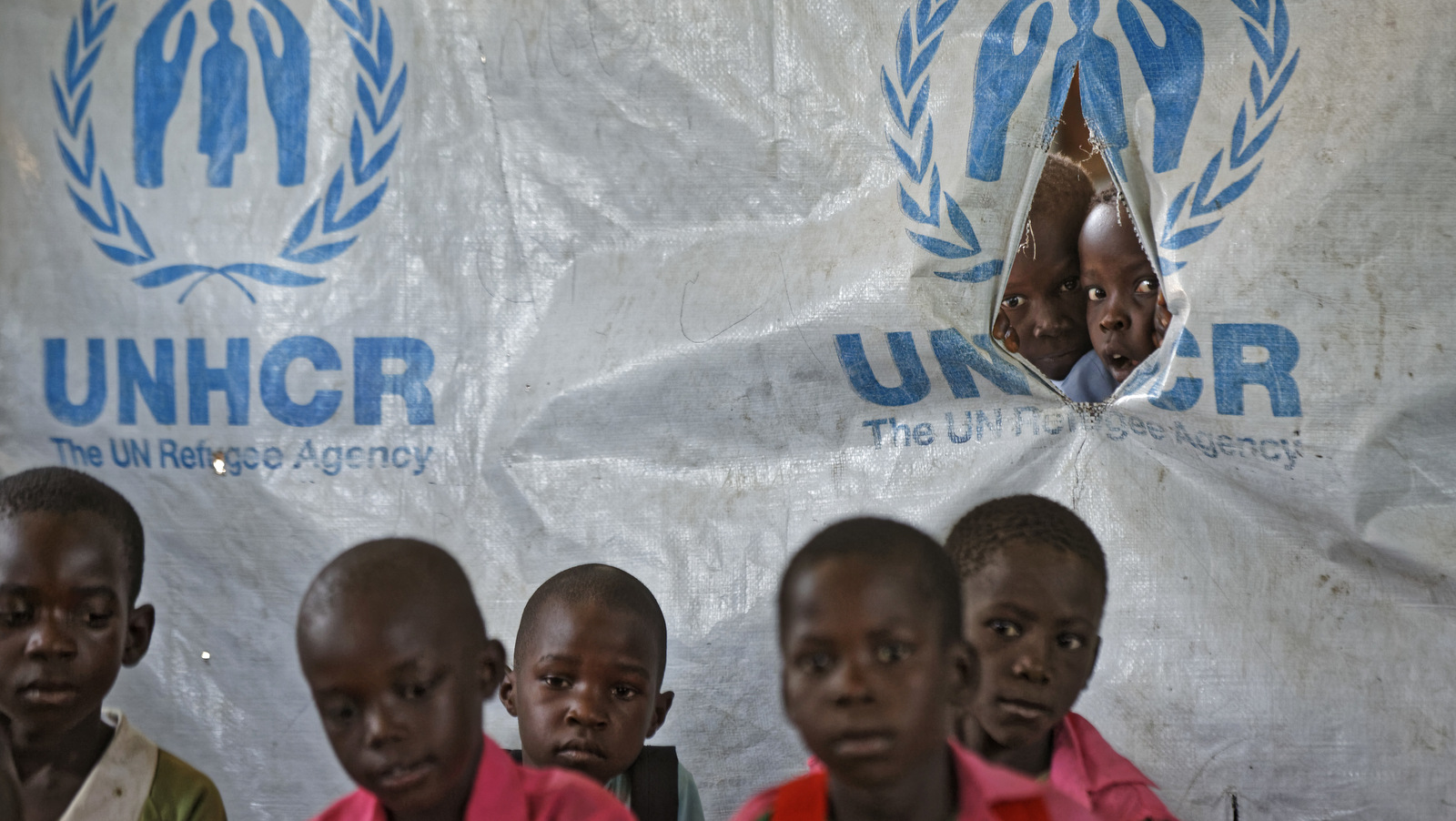 In this photo taken Wednesday, June 7, 2017, children look through a tear in the tarpaulin tents that serve as extra classrooms, for a mixed class of South Sudanese refugee children and Ugandan children, at the Ombechi Primary School in Bidi Bidi refugee settlement, northern Uganda. There is an urgent need to educate children in this overcrowded settlement that is home to over 270,000 refugees from South Sudan's civil war, despite them often having to walk long distances to the nearest school, funding shortages and a reduction in food rations for some refugees. (AP/Ben Curtis)
