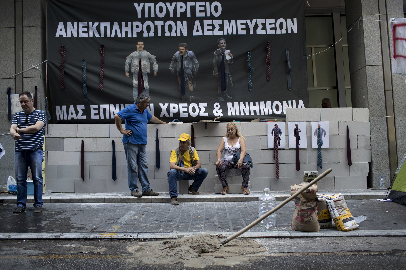 Protesting hospital staff sit in front of a wall that they built at the entrance of the Greek Finance Ministry with a banner depicting Greek Prime Minister Alexis Thipras , Deputy Health Minister Pavlos Polakis and Greek Finance Minister Euclid Tsakalotos wearing ties reading in Greek ''Ministry of broken promises" and " We drown in debt and bailouts" in central Athens. (AP/Petros Giannakouris)