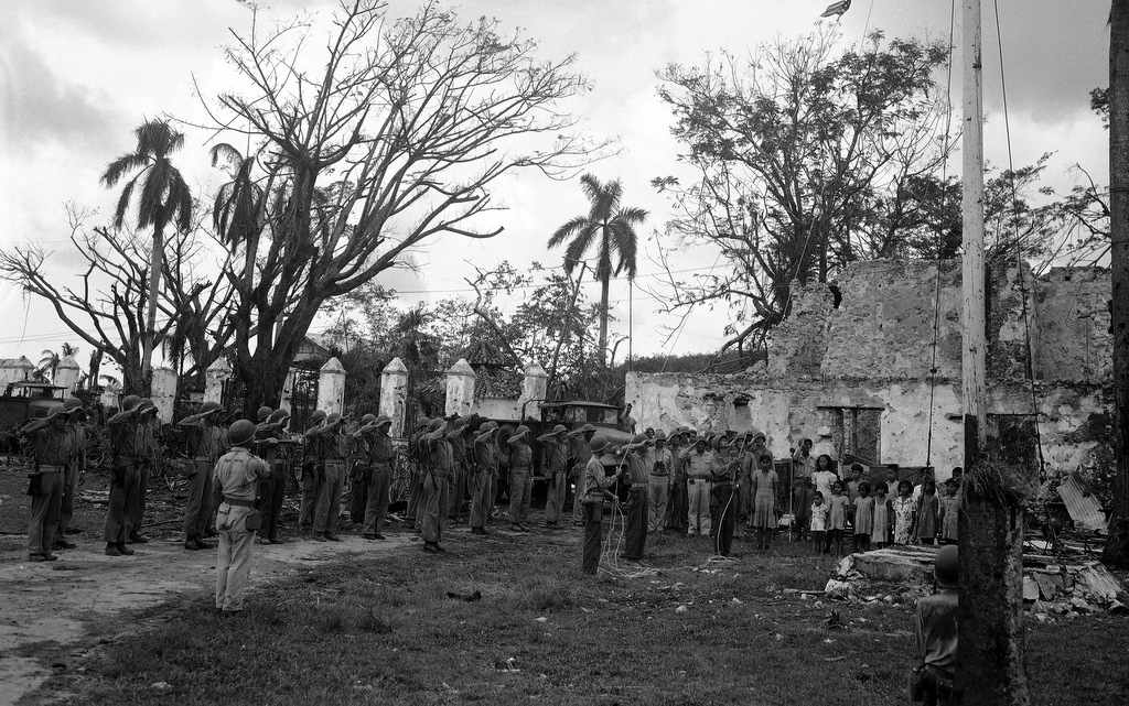 Indigenous people of Guam stand aside as American officers raise an American flag, amid the ruins of Plaza de Espana, Agana, capitol of Guam, Aug. 10, 1944. (AP/Joe Rosenthal)