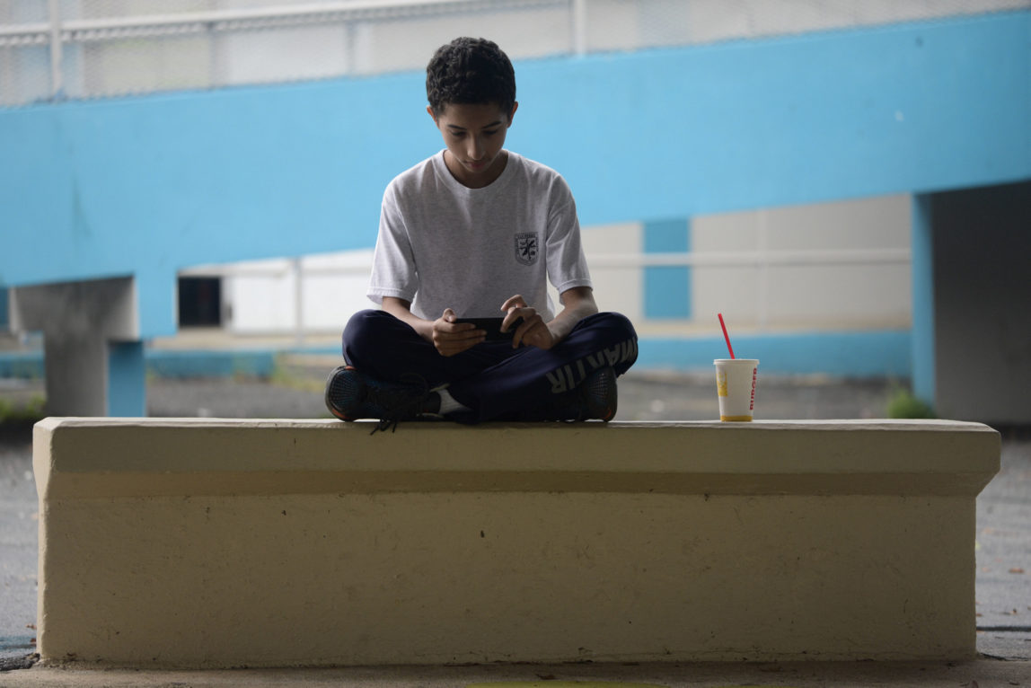 In this Friday, Oct. 13, 2017 photo, a youth sits in the courtyard of Ramon Marin Sola Elementary School, which opened its doors as a daytime community center after the passing of Hurricane Maria in Guaynabo, Puerto Rico, Most schools remain closed, leaving kids to pass the time playing on downed trees or using precious phone battery on video games, waiting for life to return to normal as the adults around them struggle to put their own lives back together. (AP Photo/Carlos Giusti)