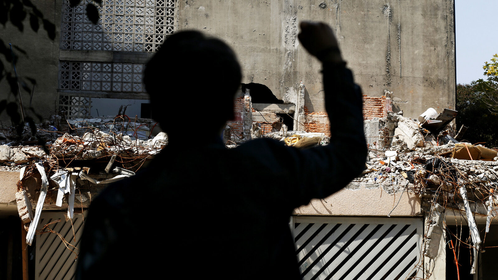 The silhouette of a man with a raised fist stands for 3 minutes of silence in front of a quake-collapsed building where several people died, on the street corner of Amsterdam and Laredo, in Mexico City, Thursday, Oct. 19, 2017. Mexico City residents gathered at different sites of quake-ravaged building one month after the Mexico 7.1 earthquake-magnitude that killed 228 people in the capital. (AP/Marco Ugarte)