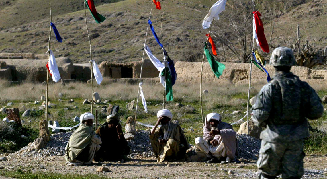A U.S soldier of 82nd Air Borne walks as villagers sit near the grave of three young boys killed in airstrike in Chinar village of Ghorak district of Kandahar province Southern Afghanistan, March 14, 2007. (AP/Rafiq Maqbool)