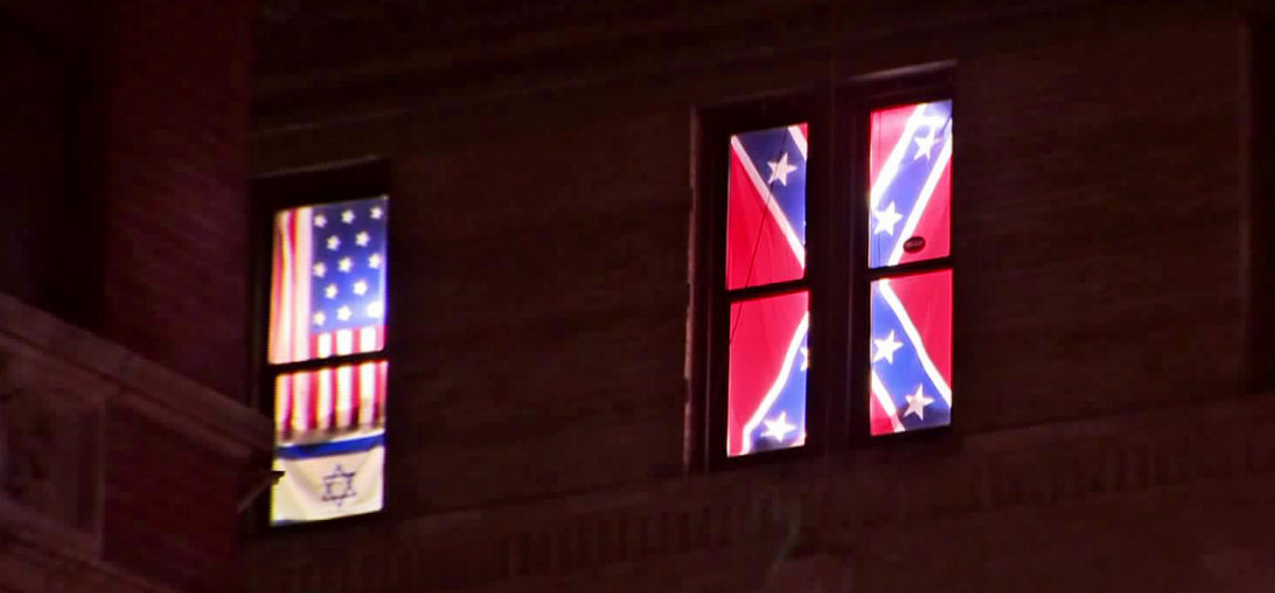 This Aug. 17, 2017 image from video shows a Confederate flag, right, displayed alongside an Israeli flag and a colonial-era American one in the seventh-floor windows of an apartment in the East Village neighborhood of New York. The flags had been there for over a year, and illuminated at night, but after an Aug. 12 white nationalist rally to preserve a Confederate statue in Charlottesville, Va., spiraled into violence the flags were met with hurled rocks, a punched-out window, a tarp hung over them and legal action before being removed. (PIX11 News via AP)