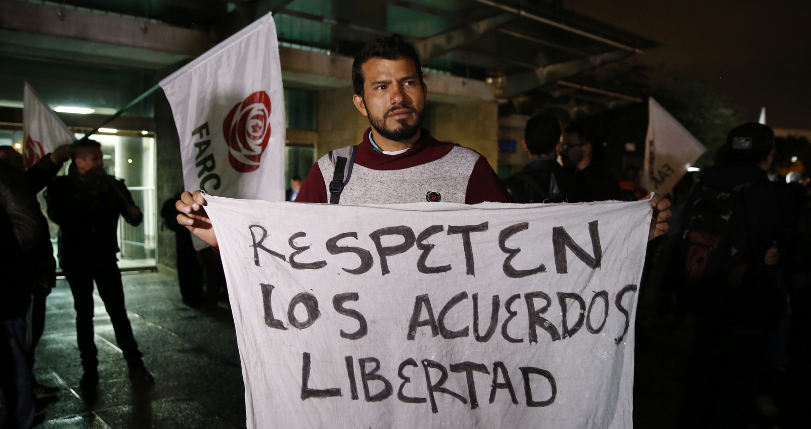 A supporter of former FARC rebel Jesus Santrich holds a sign that reads in Spanish "Respect the freedom agreements" during a protest against his arrest outside the Attorney General Office where he is being held in Bogota, Colombia, Monday, April, 9, 2018. Seuxis Hernandez, a blind rebel ideologue best known by his alias Jesus Santrich, was picked up Monday at his residence in Bogota on charges that he conspired with three others to smuggle several tons of cocaine into the U.S. with a wholesale value of $15 million. (AP Photo/Fernando Vergara)