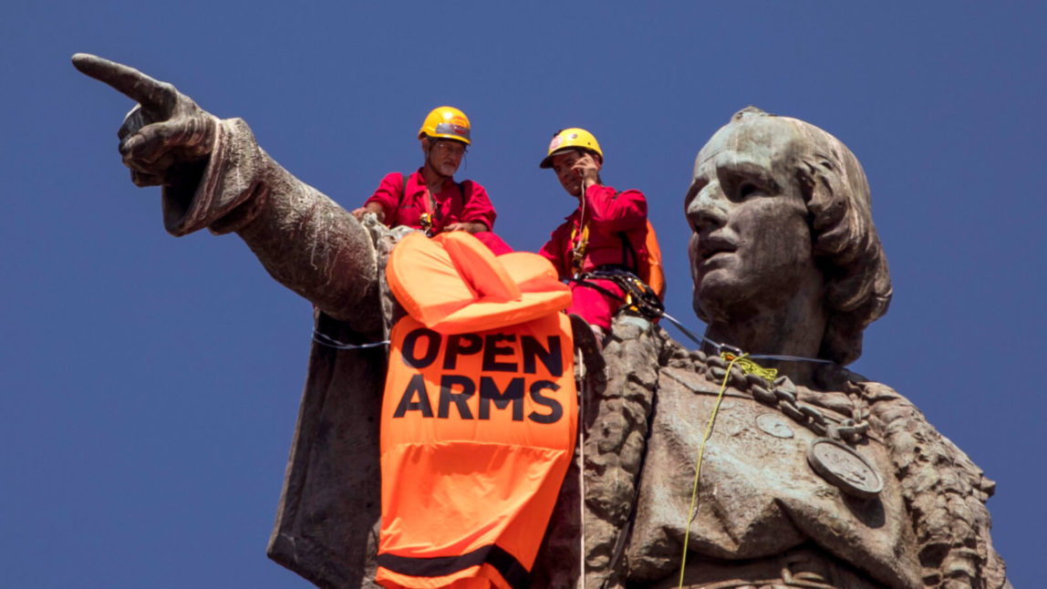 Two activists sit on top of the 60-meter (197-feet) monument Christopher Columbus tower after placing a vest with the words "Open Arms" on the statue in Barcelona, Spain, Wednesday, July 4, 2018. Activists in Barcelona have dressed in an orange life-vest a statue of 15th-century explorer Christopher Columbus to turn attention to the loss of life of migrants and refugees in the Mediterranean Sea. Emilio Morenatti | AP