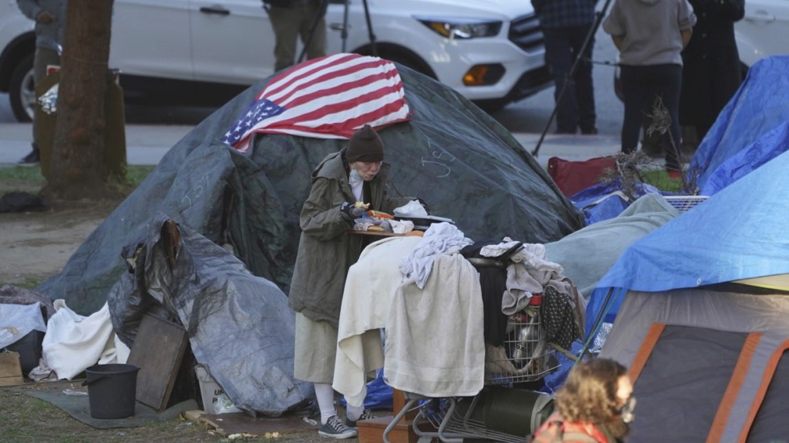 In this March 24, 2021, file photo a woman eats at her tent at the Echo Park homeless encampment at Echo Park Lake in Los Angeles. A proposal by two Los Angeles city councilmen to set up temporary camps or "tiny homes" for homeless people in beach parking lots is drawing opposition. The motion asks the city administrative officer to evaluate and identify funding for temporary sites for "single-occupancy tiny homes or safe camping" at Will Rogers State Beach in Pacific Palisades, Dockweiler Beach in Playa del Rey and Fisherman's Village in Marina del Rey. (AP Photo/Damian Dovarganes, File)