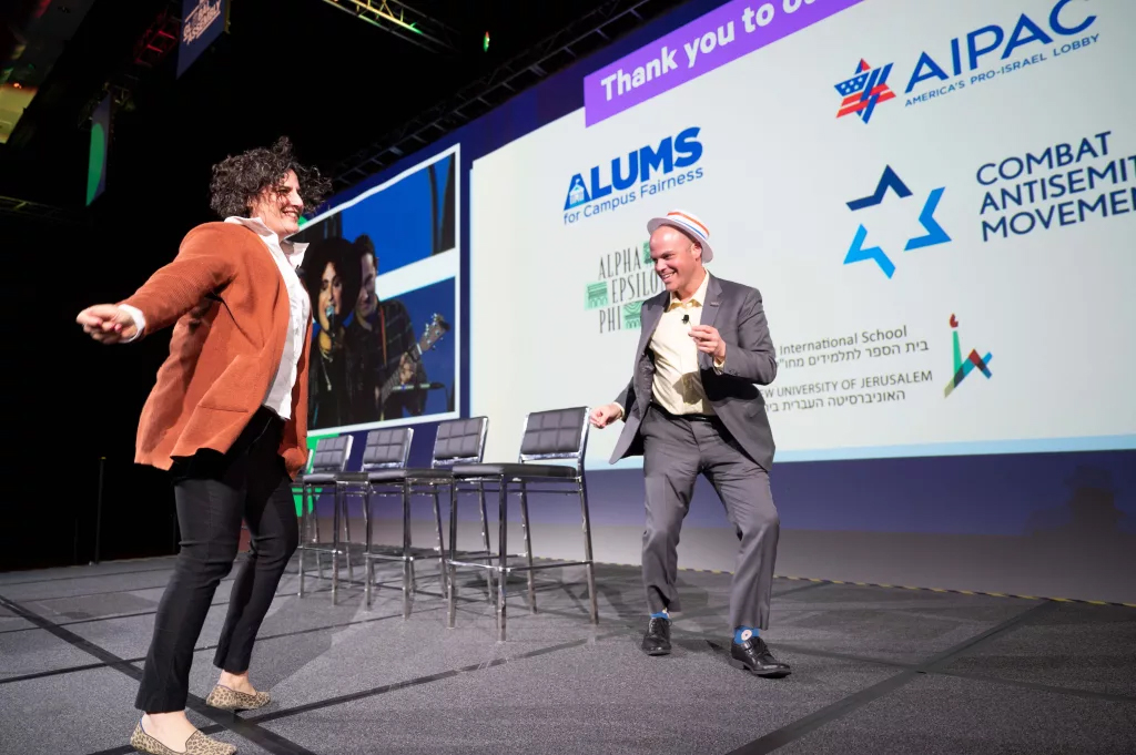 Глава GW Hillel Адена Кирштейн (слева) танцует на мероприятии Hillel 2022 года. На заднем плане виден логотип AIPAC, противоречивого произраильского лобби.